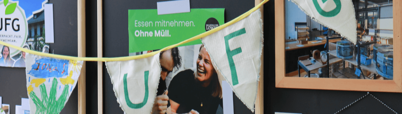 eine selbstgebastelte Wimpelkette mit den Buchstaben U, F, G hängt vor einer schwarzen Aufstellwand, auf der ein Plakat zur Müllvermeidung hängt und ein paar Bilder in Rahmen vom Aufbau des Ladens