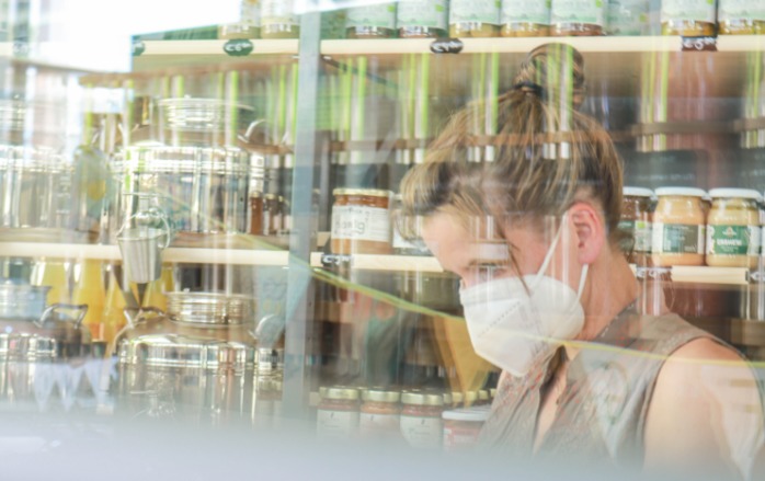 Durchs Fenster sieht man eine Besucherin mit vorgeschriebenem Mund-Nasen-Schutz vor einem Waren-Regal