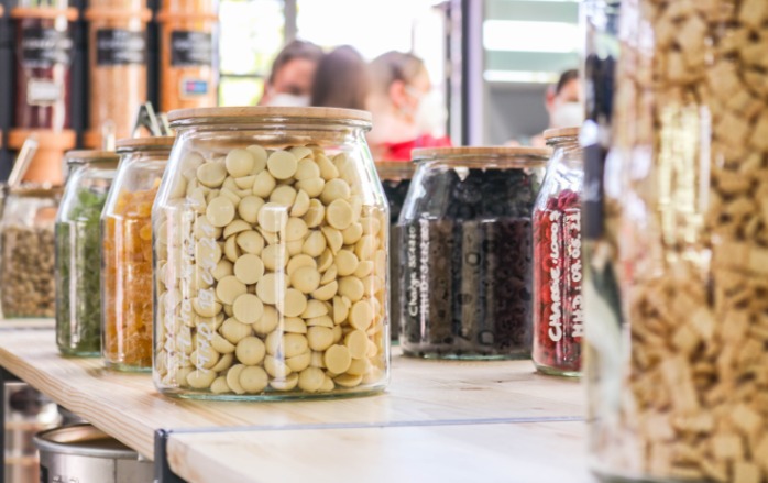 Im Vordergrund befinden sich große, handbeschriftete Glasbehälter mit z. B. weißen Schokodrops gefüllt. Dahinter stehen Besucher bei der Eröffnungsfeier im Laden, alle tragen den vorgeschriebenen Mund-Nasen-Schutz