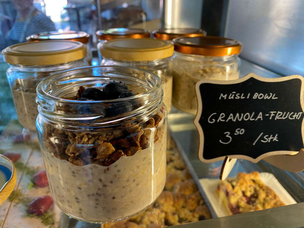 Müsli-Bowl mit einer Granola mit Heidelbeeren angerichtet in einem Schraubglas in der Verkaufsvitrine des Ladens. Daneben steht ein kleines handbemaltes Schild mit der Aufschrift: "Müsli Bowl Granola Frucht 3,50/Stück