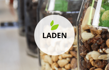 Ausschnitt mehrerer nebeneinander stehender Glasgefäße, die mit Nüssen und Studentenfutter gefüllt sind. Darüber mittig ein transparenter weißer Kreis mit dem Begriff LADEN. Darüber befinden sich zwei kleine grafische grüne Blätter.