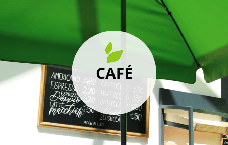 Eine schwarze Tafel mit goldenem Rahmen auf dem sehr kunstvoll die Kaffeekarte von Hand darauf geschrieben ist. Davor steht ein großer grüner Schirm. Eine Ecke des Gebäckregals ist noch zu erkennen. Mittig darauf platziert befindet sich ein transparenter weißer Kreis mit schwarzer Schrift. Der Begriff lautet CAFÉ. Darüber befinden sich zwei kleine grüne grafische Blätter.