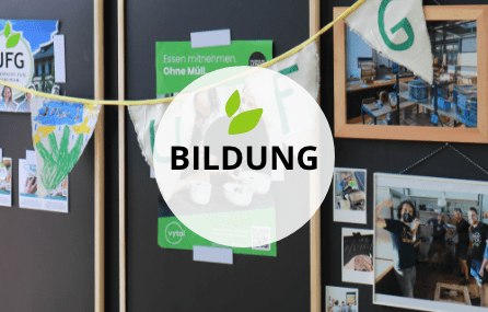 Schwarze Stellwände mit handgefertigtem Wimpel, der mit dem Buchstaben U, F, G und einer Kinderzeichnung verziert ist. Dahinter befindet sich ein Poster zur Müllvermeidung und ausgedruckte Bilder, die den Ladenaufbau zeigen. Darüber ist ein transparenter weißer Kreis auf dem mit schwarzer Schrift der Begriff BILDUNG steht. Darüber sind zwei kleine grüne grafische Blätter.