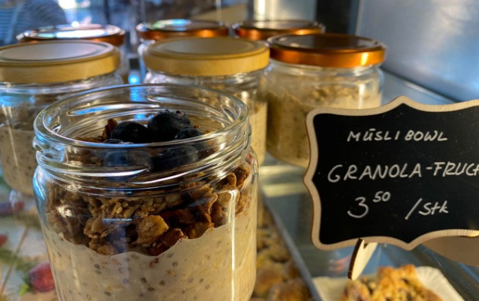 Appetitlich angerichtete Granola mit Frucht in einem Glas mit offenem Deckel. Daneben ein handbemaltes Schild mit dem Preis 3,50 € pro Stück und die Bezeichnung "Granola-Frucht-Müsli-Bowl"