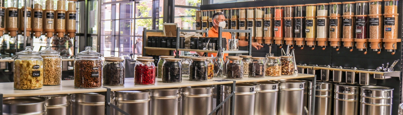 Ansicht der gFrisch gefüllte Regale Unverpackt-Laden. Mittendrin steht eine männliche Person in einem orangenen T-Shirt.